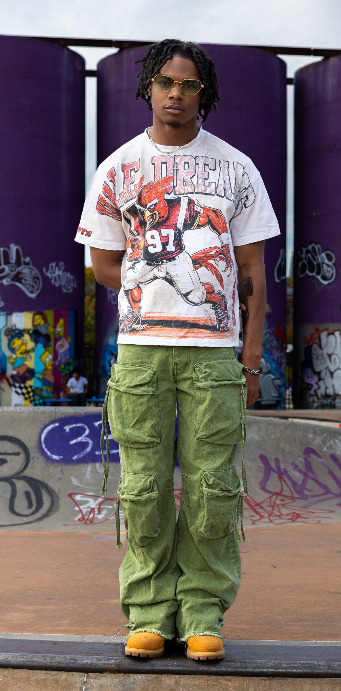 Person wearing a graphic t-shirt and green cargo pants standing in front of a graffiti-covered wall.