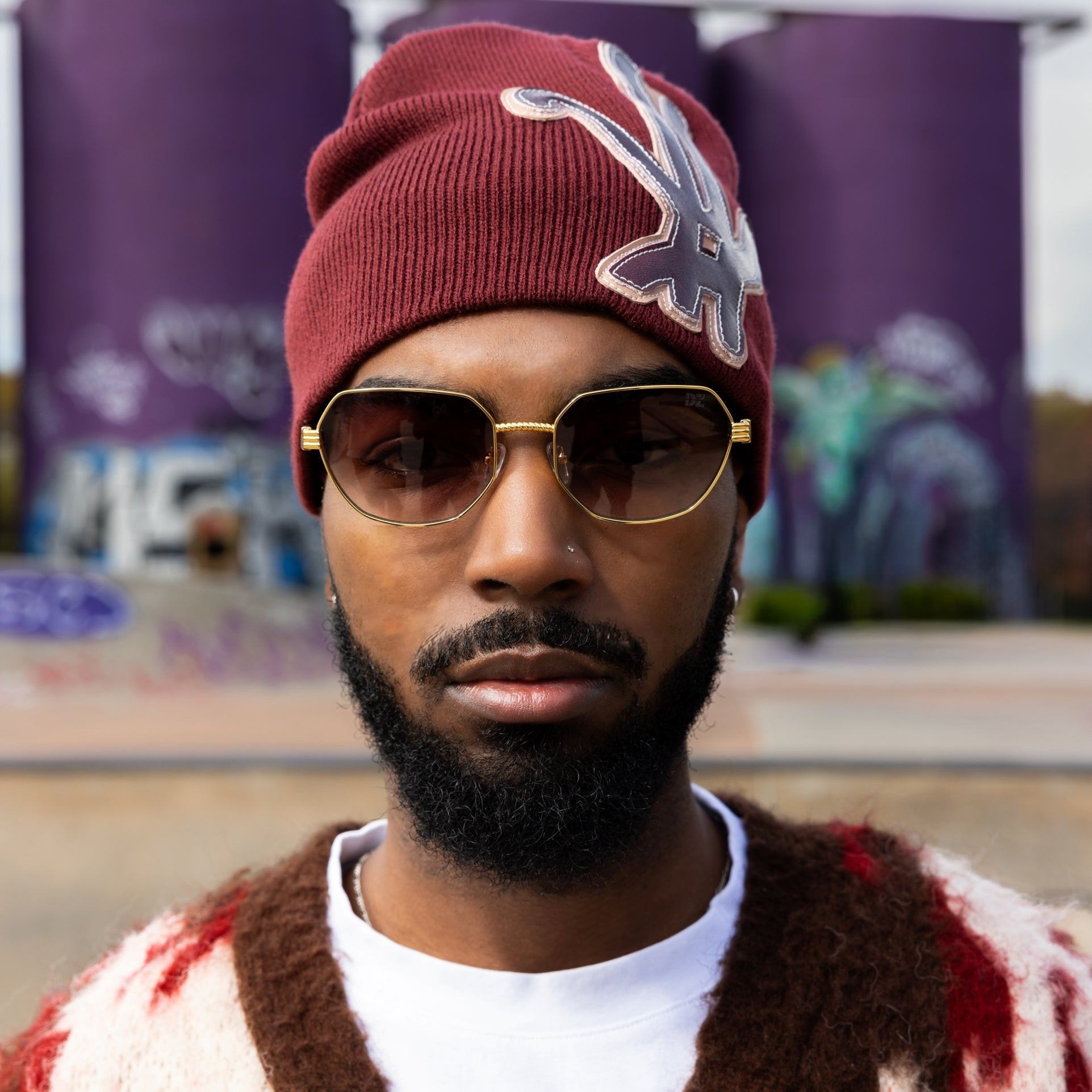 Man wearing a maroon beanie with a logo, sunglasses, and a patterned sweater in an urban setting.