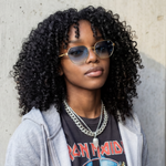 Person with curly hair wearing sunglasses and a graphic t-shirt against a concrete wall.