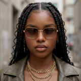 Woman wearing gold sunglasses and multiple necklaces with a blurred street background