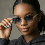 A close-up of a woman wearing oversized square gradient sunglasses with gold metal frames and a small 'DZ' logo on the temple, holding one lens with her hand.