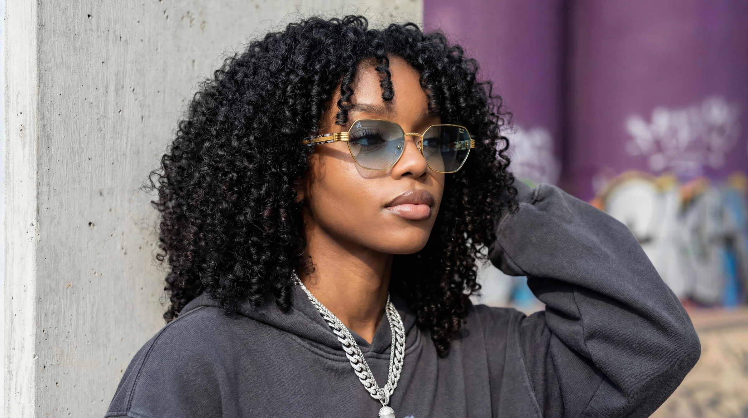 Woman with curly hair wearing sunglasses and a dark hoodie against a blurred background