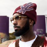 A close-up, outdoor photo of a man wearing a maroon knit beanie with a large, stylized leather appliqué design, gold-rimmed hexagonal sunglasses with tinted lenses, a small silver hoop earring, and a shaggy brown, white, and red patterned cardigan over a white t-shirt. He has a neatly trimmed beard and is looking off to the left. In the background, there are blurred, tall purple concrete structures with graffiti visible at the base.